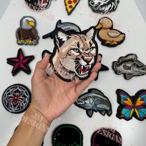 May include: A selection of embroidered patches with diverse designs, such as a cougar, wolf, pizza slice, eagle, duck, fish, butterfly, spider, and skulls. The patches are in various colours and styles, with the JM Designs logo visible.