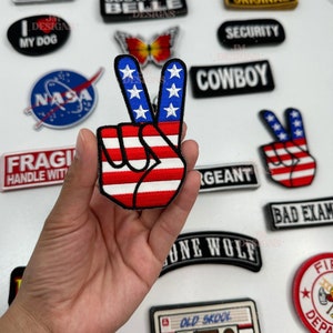 May include: A red, white, and blue peace sign patch with a hand making the peace sign. The patch is surrounded by other patches with various designs, including a butterfly, a cowboy hat, and a cassette tape.