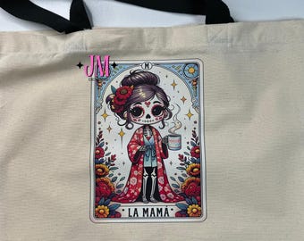 Puede incluir: Bolso tote color crema con asas negras. El bolso presenta una colorida ilustración de una mujer esqueleto sosteniendo una taza, con el texto "LA MAMA" en la parte inferior. La ilustración está enmarcada con detalles florales y de estrellas.