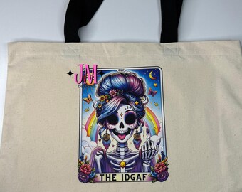 Puede incluir: Bolso tote de lona beige con asas negras. La parte delantera presenta una colorida ilustración de una mujer esqueleto con cabello azul y morado, un arcoíris y el texto "THE IDGAF". El esqueleto está mostrando el dedo medio.