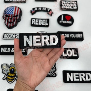May include: A hand holding a black and white embroidered patch that says "NERD" in white letters. The patch is surrounded by other patches with various designs and text, including "I Make Cute Babies", "Good Vibes Only", "Rebel", "Army", "No Thanks", "Trouble Maker", "Wild & Free", "Did You Die?", "LOL", "Rock Star", "My Soldier", "Biker Babe", and "Nerd".