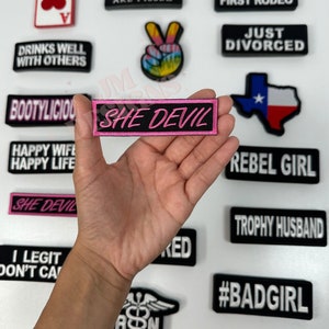 以下が含まれることがあります： 「SHE DEVIL」というピンクの文字が書かれた黒とピンクのアイロン接着パッチを手に持っている。パッチは、「We Be People Are Pissed」、「This Is My First Rodeo」、「Just Divorced」、「Rebel Girl」、「Trophy Husband」、「#BADGIRL」など、さまざまなフレーズやデザインのアイロン接着パッチに囲まれている。