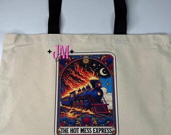 Puede incluir: Bolso tote color crema con asas negras. Presenta un gráfico colorido de un tren azul en llamas, con el texto "THE HOT MESS EXPRESS". El diseño incluye rosas, una luna creciente y estrellas.