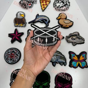 May include: A black and white embroidered patch of a drum with drumsticks crossed over it. The patch is surrounded by other embroidered patches, including a wolf, a duck, a fish, a skull, a spider, a butterfly, a pizza slice, a star, a marlin, and an eagle.
