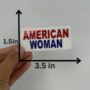 May include: White iron-on patch with red and blue embroidered text that reads "AMERICAN WOMAN". The patch measures 3.5 inches wide and 1.5 inches tall.
