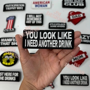 May include: A black and white embroidered patch with the text "You look like I need another drink". The patch is held in a hand against a white background with other patches visible.