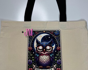 Puede incluir: Bolso tote color crema con asas negras. El bolso presenta un gráfico colorido de un búho con los ojos cerrados, una luna creciente y flores. El texto "THE NIGHT OWL" está en una pancarta debajo del búho.