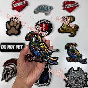 May include: A collection of embroidered iron-on patches featuring a variety of designs, including a parrot, a dog paw print, a snake, a turtle, a heart with the word "Grandma" on it, a heart with the word "Grandpa" on it, a service dog symbol, a helmet, a dog head, and a sign that says "Do Not Pet".