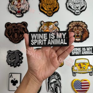 Puede incluir: Un parche bordado en blanco y negro con el texto "Wine is my spirit animal". El parche está rodeado de otros parches bordados con animales, incluyendo tigres, un oso, un león, un bulldog, un casco espartano, un coche amarillo y una bandera estadounidense en forma de corazón.