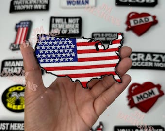 Può includere: Una toppa ricamata di colore rosso, bianco e blu a forma degli Stati Uniti d'America. La toppa è tenuta in una mano.