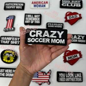 May include: A collection of iron-on patches with various designs and sayings. The patches are black, white, red, blue, and yellow. Some of the patches include the text "Crazy Soccer Mom", "Participation Trophy Wife", "American Woman", "Mid Day Drinking Club", "Dad", "I Will Not Remember Any Of This", "It's 5 O'Clock Somewhere", "Manifest That Shit", "Just Here For The Drink", "You Look Like I Need Another Drink", and "Mom".