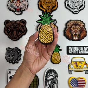 Puede incluir: Una colección de parches bordados para planchar con varios diseños, que incluyen una piña, un tigre, un león, un oso, un bulldog, un coche, un corazón con la bandera estadounidense, un casco espartano, una granada y un as de picas.