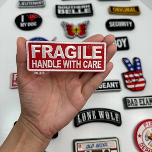 May include: A red and white embroidered patch with the words "FRAGILE HANDLE WITH CARE" in white lettering. The patch is held in a hand.