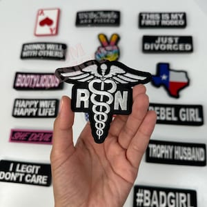 Puede incluir: Un parche termoadhesivo negro y blanco con un símbolo de caduceo y las letras "RN" en blanco. El parche está siendo sostenido por una mano.