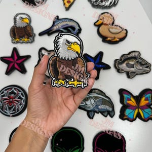 Puede incluir: Una colección de parches bordados con varios diseños, incluyendo un águila calva, un lobo, una porción de pizza y una mariposa. Los parches son de varios colores y formas, algunos mostrando el texto "JM Designs".
