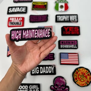 May include: A collection of embroidered iron-on patches with various phrases and designs. The patches are black with white and pink lettering. Some of the phrases include "High Maintenance", "Savage", "Sweetheart", "Trophy Wife", "Yes I Do Bite", "Warning Bombshell", "Big Daddy", "Good Girl Gone Bad".