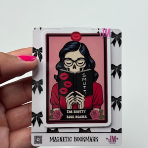 May include: A magnetic bookmark featuring a skull-faced woman reading a book titled "SMUTT." The woman wears glasses and a red top. The bookmark has a pink background with a red lip design and the text "THE SMUTTY BOOK READER."