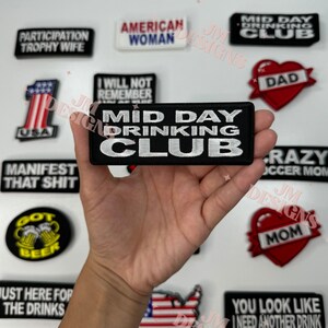 May include: A black and white embroidered patch with the text "Mid Day Drinking Club". The patch is held in a hand against a background of other patches with various designs and text.