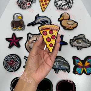 May include: A collection of embroidered iron-on patches featuring various designs, including a pizza slice, a skull, a butterfly, a fish, a duck, a wolf, a star, a spider, and an eagle.