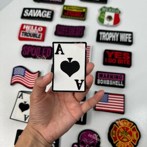 May include: A collection of iron-on patches featuring various designs, including a playing card, an American flag, a skull and crossbones, a fire department logo, and text patches with phrases like "Savage", "MILF", "Trophy Wife", and "Warning Bombshell".