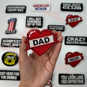May include: A collection of iron-on patches with various designs and sayings. The patches include "Participation Trophy Wife", "American Woman", "Mid Day Drinking Club", "I Will Not Remember Any Of This", "It's 5 O'Clock Somewhere", "Dad" in a red heart, "Manifest That Shit", "Crazy Soccer Mom", "Mom" in a red heart, "Just Here For The Drinks", and "You Look Like I Need Another Drink".