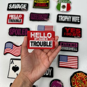 Puede incluir: Una colección de parches bordados para planchar con diferentes frases y diseños. Los parches son rojos, negros, blancos, rosas y azules. Algunas de las frases en los parches incluyen "Hello My Name Is Trouble", "Savage", "Milf", "Trophy Wife", "Warning Bombshell" y "Yes I Do Bite".