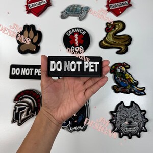 May include: A collection of embroidered iron-on patches featuring various designs, including a paw print, a snake, a turtle, a heart with the word "GRANDPA" on it, a heart with the word "GRANDMA" on it, a service dog patch, a parrot, a roman helmet, a bulldog, and a patch that says "DO NOT PET".