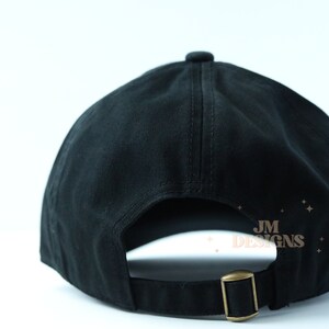 May include: Black baseball cap with an adjustable strap and a small gold logo that says "JM DESIGNS" on the back.