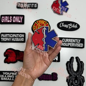 May include: A collection of embroidered patches with various designs and text. Patches include an emergency medical services symbol, a fire department emblem, and phrases like "Girls Only" and "Currently Unsupervised."