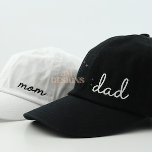 May include: Two baseball caps, one white with the word "mom" embroidered in black, and one black with the word "dad" embroidered in white.