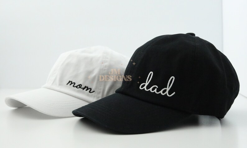 May include: Two baseball caps, one white with the word "mom" embroidered in black, and one black with the word "dad" embroidered in white.