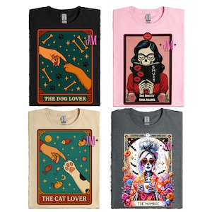 May include: Four t-shirts with tarot card-style designs. The shirts are black, pink, tan, and gray. Designs include "The Dog Lover", "The Shutty Book Reader", "The Cat Lover", and "The Mombie". Each design features unique illustrations and text.