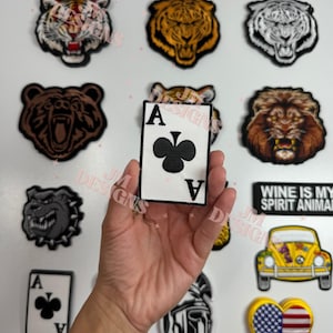 May include: A close-up of a hand holding a white patch with a black ace of clubs playing card design. The patch is surrounded by other patches, including a tiger, a bear, a lion, a bulldog, a car, a heart with the American flag, and a gladiator.