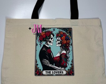 Puede incluir: Bolso tote color crema con asas negras. Presenta un diseño de carta del tarot con dos esqueletos adornados con flores, mirándose. La carta dice "THE LOVERS" en un marco decorativo. El bolso está hecho de un material de lona resistente.