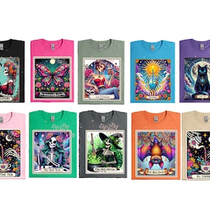 May include: Ten different t-shirts featuring colorful illustrations of skeletons, butterflies, cats, and other imagery. The shirts are all in different colors, including pink, blue, green, and black. The text on the shirts includes "The Lovers", "La Tosca", "The Ritual Butterfly", "The Book", "The Cat", "Spill The Tea", "The Shit Trick", "Bot That Crazy", "The Shit Stirrer", and "El Chisme".