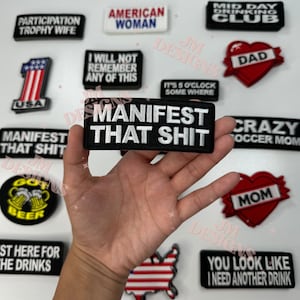 May include: A collection of iron-on patches with various designs and sayings. The patches include "Participation Trophy Wife", "American Woman", "I Will Not Remember Any Of This", "Mid Day Drinking Club", "Dad", "It's 5 O'Clock Somewhere", "Manifest That Shit", "Crazy Soccer Mom", "Got Beer", "Mom", "St Here For The Drinks", "You Look Like I Need Another Drink", and "Manifest That Shit".