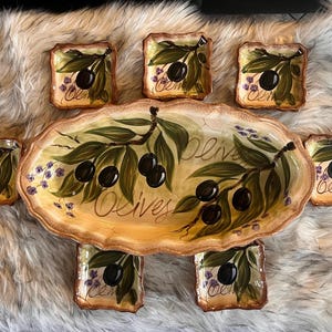 May include: A hand-painted ceramic serving set featuring an oval platter and six square dishes. The design includes olive branches with black olives and the word "Olives" in script. The set has a rustic, Mediterranean style.