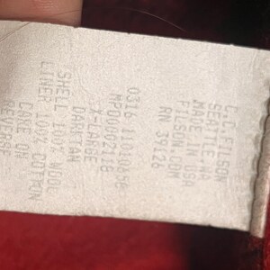 Puede incluir: Una etiqueta blanca con texto negro, parcialmente oscurecida por un dedo. La etiqueta dice "C.C. FILSON SEATTLE WA MADE IN USA FILSON.COM RN 39126". El texto adicional incluye "0316 11010858 MP00002118 X-LARGE DARKTAN SHELL 100% WOOL LINER 100% COTTON CARE ON REVERSE".
