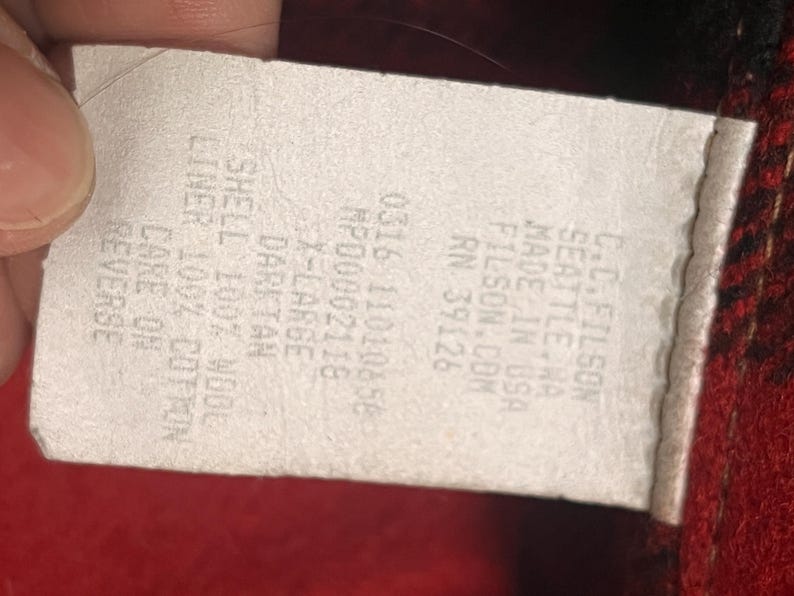 Puede incluir: Una etiqueta blanca con texto negro, parcialmente oscurecida por un dedo. La etiqueta dice "C.C. FILSON SEATTLE WA MADE IN USA FILSON.COM RN 39126". El texto adicional incluye "0316 11010858 MP00002118 X-LARGE DARKTAN SHELL 100% WOOL LINER 100% COTTON CARE ON REVERSE".