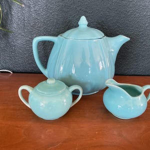 May include: A light blue ceramic tea set consisting of a teapot with a lid, a lidded sugar bowl with two handles, and a creamer pitcher with one handle. Each piece features a smooth, glossy finish and a rounded, bulbous shape with vertical ridges.
