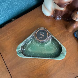 May include: Green ceramic ashtray with a triangular shape and a central circular area. The ashtray has a speckled finish and a dark brown border. A decorative emblem with the letter "S" is in the center.