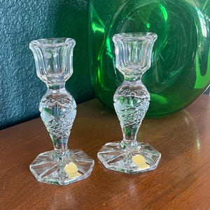 May include: Two clear glass candle holders with a cut glass design. The candle holders have a hexagonal base and a round top. The candle holders are on a wooden surface.