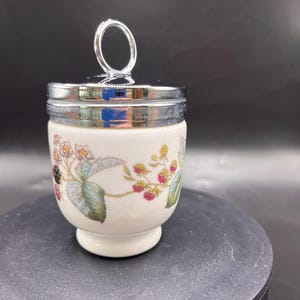 May include: A white ceramic egg cup with a silver metal lid and handle. The cup is decorated with a floral pattern featuring red berries, green leaves, and small yellow flowers. The egg cup sits on a small, round base.