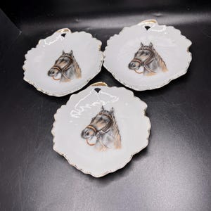 May include: Three white, leaf-shaped decorative dishes with a gold trim. Each dish features a detailed painting of a horse's head with a bridle. The dishes are arranged on a dark surface.