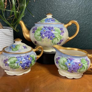 May include: A porcelain tea set featuring a teapot with a lid, a sugar bowl with a lid, and a creamer. Each piece is decorated with hand-painted purple violet flowers and green leaves on a pale blue background, accented with gold handles and trim.
