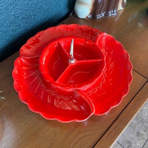 May include: A bright red, vintage divided serving dish. The dish has a central section with three compartments and a larger, petal-shaped outer section. A silver metal handle is in the center. The dish is made of ceramic or porcelain.