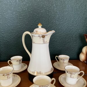 May include: A white and beige tea set with gold accents. The set includes a tall coffee pot, six teacups, and saucers. The design features floral and geometric patterns. The set is arranged on a wooden surface against a dark green background.