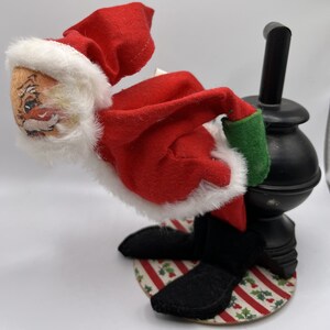 May include: A plush Santa Claus doll in a red suit with white trim, a white beard, and black boots. He is leaning into a black chimney with a green cuff on his left arm. The doll is standing on a round, white and red striped base with green holly leaves.