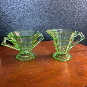 May include: Two matching, vintage, light green glass cream and sugar set. Each piece has a geometric design with a handle and sits on a hexagonal base. The set is displayed on a wooden surface.