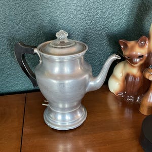 May include: A vintage silver-colored electric coffee pot with a black handle and a clear glass lid. The coffee pot has a spout and sits on a wooden surface. A ceramic cat figurine is visible in the background.